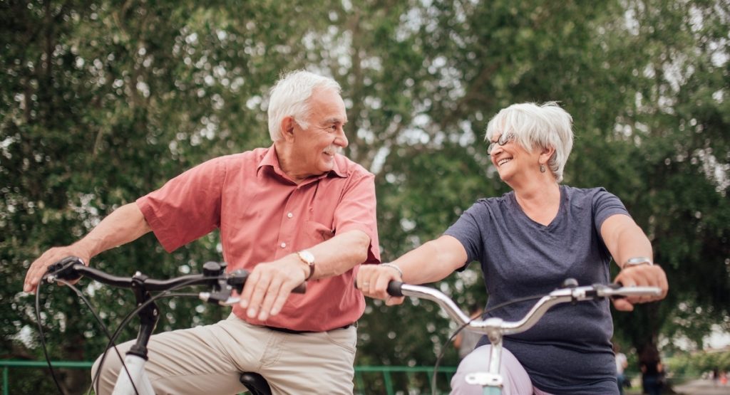 Φυσιοθεραπεία για ηλικιωμένους Ισορροπία και πρόληψη πτώσεων. Η εικόνα δείχνει δύο χαρούμενους ηλικιωμένους που κάνουν ποδήλατο.
